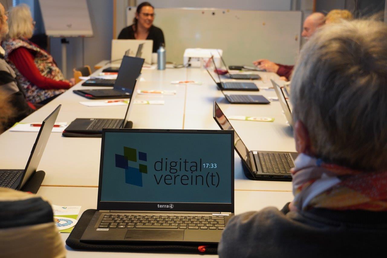 Menschen sitzen rund um einen großen Arbeitstisch, vor ihnen stehen Laptops.